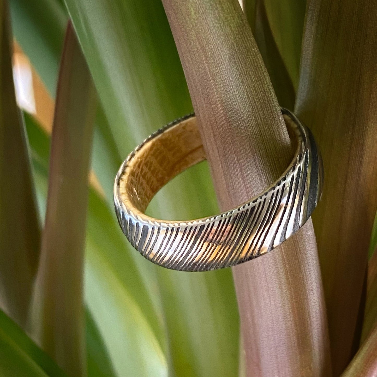 Photo of Damascus Steel Acid Finish Band with Whiskey Barrel Wood Interior Inlay in a decorative scene, designed by , available at Plumb Gold & Plumb Silver