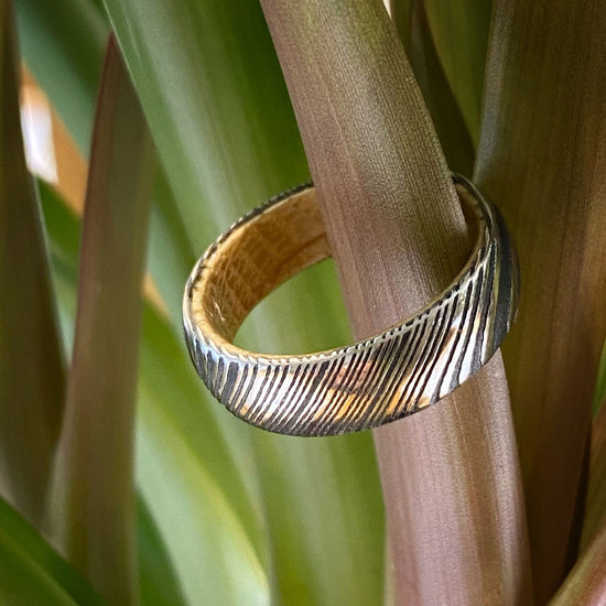 Photo of Damascus Steel Acid Finish Band with Whiskey Barrel Wood Interior Inlay in a decorative scene, designed by , available at Plumb Gold & Plumb Silver